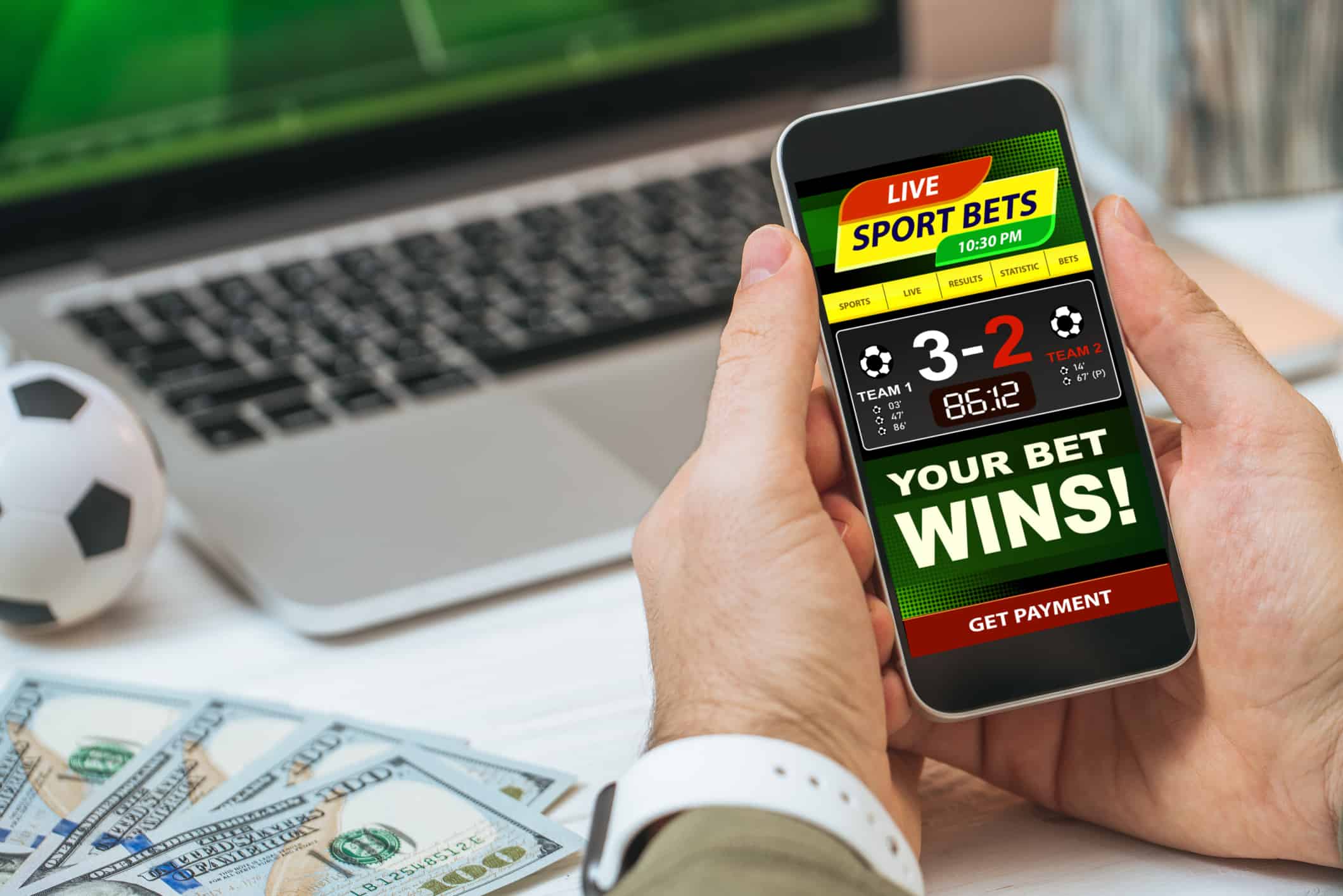 Cropped shot of male hands holding smartphone with bookmaker's mobile application interface design template. Man making bets while watching soccer play live broadcast online on his laptop.