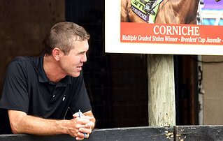 Tristan de Meric leans forward against a rail in front of horse stalls, holding a pen in his hands while viewing (presumably) horses passing by. He’s standing next to a photo of a horse that graduated from his program, Corniche. The sign further describes Corniche as a Multiple Graded Stakes winner and winner of the 2021 Breeders Cup Juvenile (G1). (Photo: ©Judit Seipert)