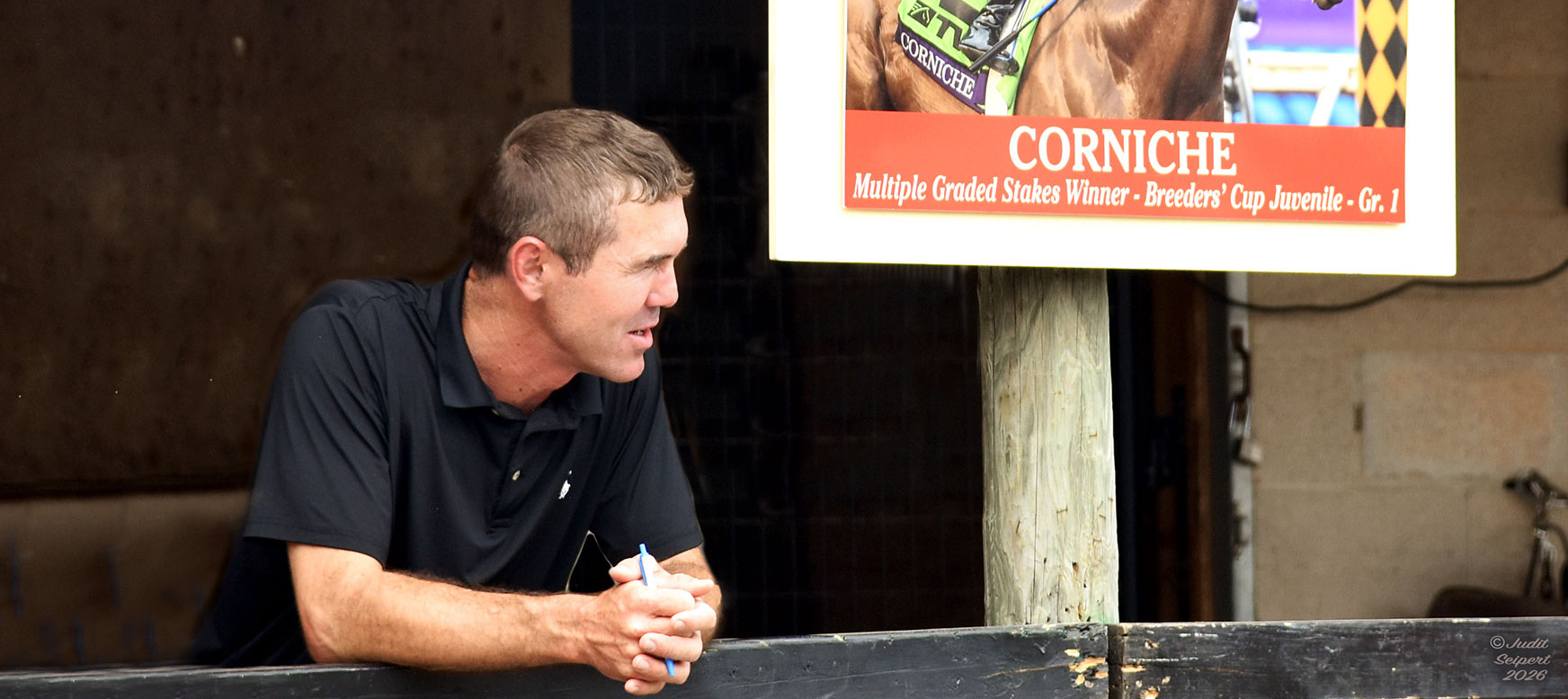 Tristan de Meric leans forward against a rail in front of horse stalls, holding a pen in his hands while viewing (presumably) horses passing by. He’s standing next to a photo of a horse that graduated from his program, Corniche. The sign further describes Corniche as a Multiple Graded Stakes winner and winner of the 2021 Breeders Cup Juvenile (G1). (Photo: ©Judit Seipert)