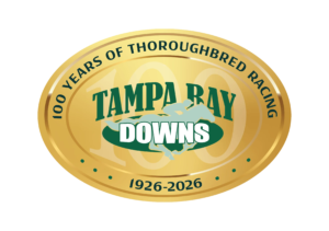The 100 years anniversary logo for Tampa Bay Downs. The logo is a golden, oval emblem, with thin outlines surrounding the circumference of the emblem that make the vector look like shining gold. Dark green curving text along the outside of the logo reads, “100 Years of Thoroughbred Racing, 1926 - 2026.” In the middle of the logo, Tampa Bay Downs standard brand logo (consisting of a galloping racehorse and jockey, overlapping the name “Tampa Bay Downs” in dark and light green) is centered overtop a large “100,” which is partially transparent and fades into the gold of the logo.