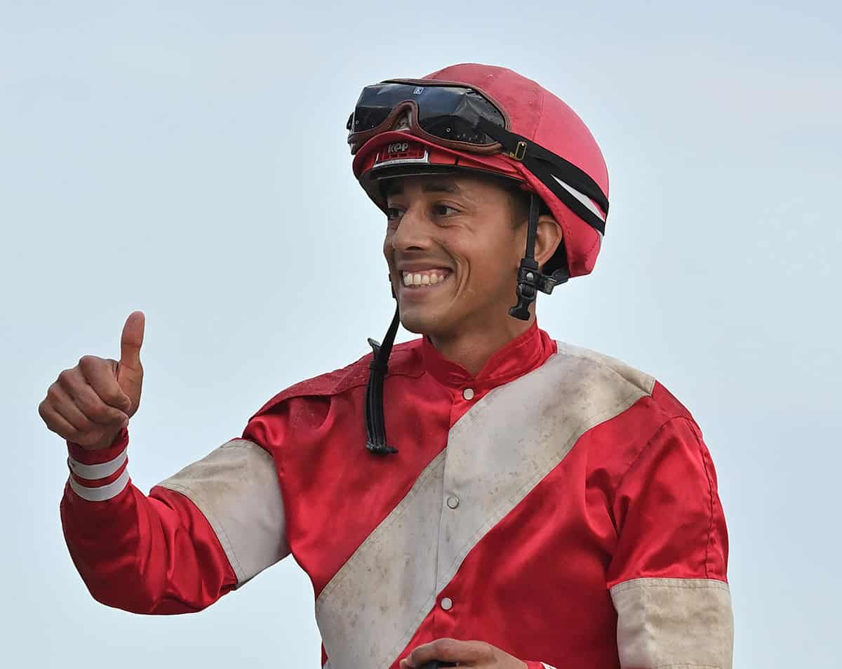 Jockey Edgard Zayas, dressed in red silks with a large white stripe that runs diagonal across his chest, and two large bands of white on either arm, smiles at someone out of frame, giving them a victorious thumbs up while astride a racehorse (also out of frame). (Photo: ©Lauren King)