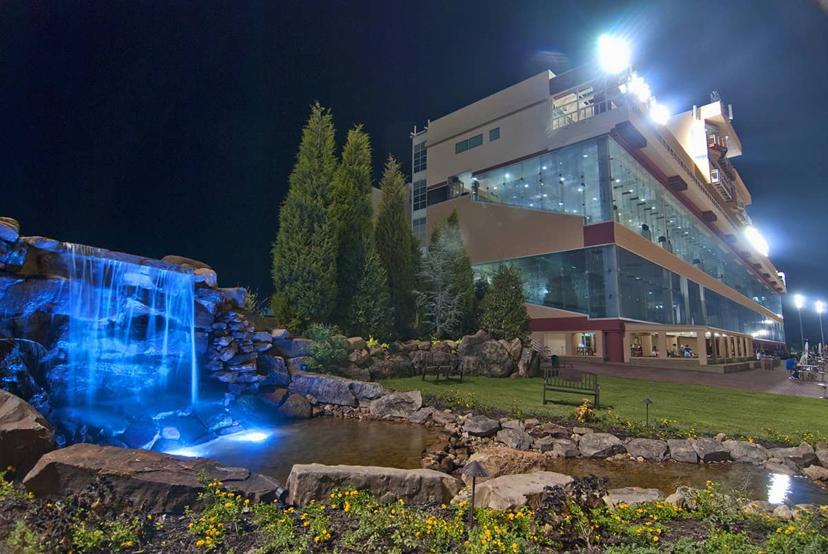 The Remington Park grandstands lit up at night. Next to the grandstands, a water feature consisting of a waterfall, lit up in glowing blue lights, flows into a small pond. (Photo: ©Dustin Orona)