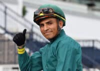 Jockey Renzo Rojas dressed, in blue-green silks with yellow accents, smiles at the camera and sends a thumbs up to the photographer. He is presumably sitting on a racehorse who is out of frame of the camera. (Photo: ©Coglianese)
