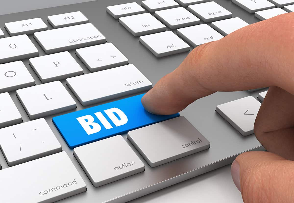 Close up of a keyboard. The keyboard’s “shift” key has been replaced with a blue key that says “Bid” in all capital letters. A finger is pressing “bid.” (Photo: ©frender)