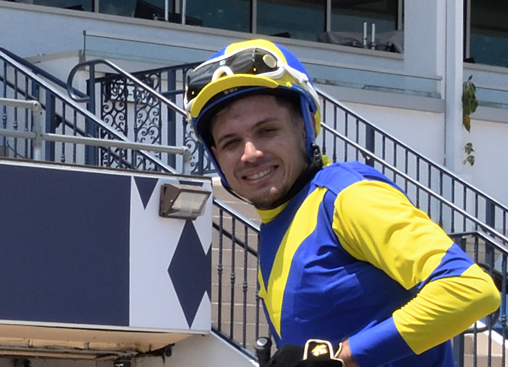 Jockey Cipriano Gil turns to face the camera, smiling while riding a horse trackside. (Photo: ©Adam Coglianese)