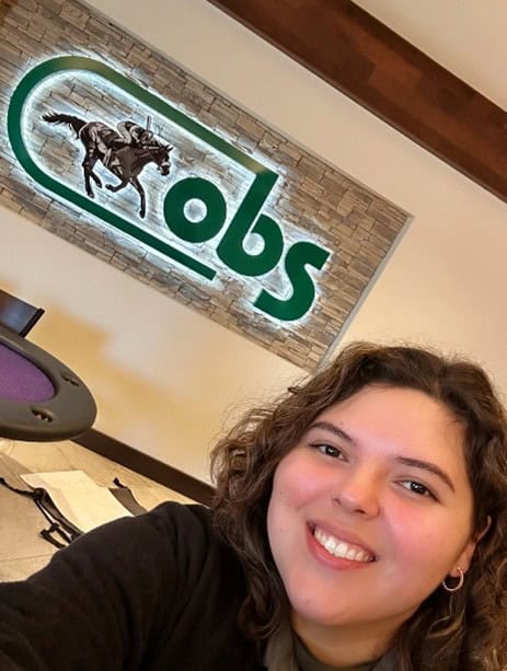 Photo of FTBOA Intern Rebecca Ramirez smiling while taking a selfie. She stands in front of the logo sign for OBS (Ocala Breeders’ Sales Company), which is lit from behind, while visiting the OBS auction grounds. (Photo Courtesy: Rebecca Ramirez)