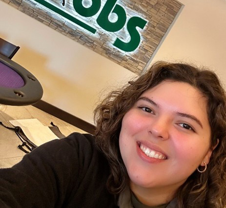 Photo of FTBOA Intern Rebecca Ramirez smiling while taking a selfie. She stands in front of the logo sign for OBS (Ocala Breeders’ Sales Company), which is lit from behind, while visiting the OBS auction grounds. (Photo Courtesy: Rebecca Ramirez)