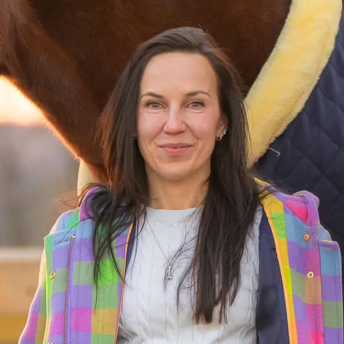 Cropped headshot of Jessica Paquette, smiling for the camera as she stands against her horse’s neck. The lighting is golden from a presumed sunset, and the horse’s face is out of frame. Both are dressed for cold weather, with the horse wearing a thick navy blanket, while Paquette wears a checkered pastel jacket. (Photo Courtesy: Jessica Paquette)