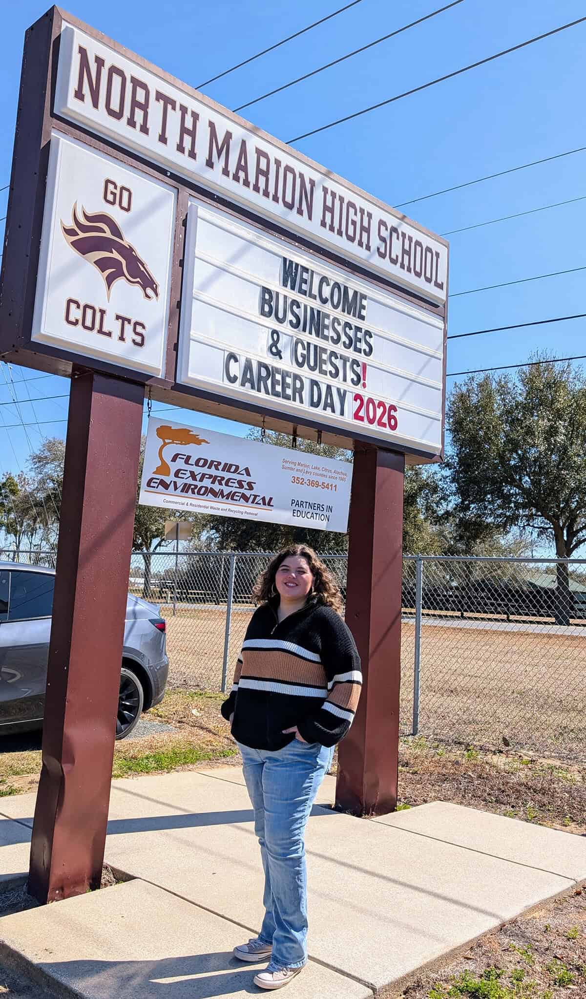 Rebecca Ramirez smiles for a photo, standing beneath a sign for the North Marion High School during their 2026 Career Fair. (Photo: ©Tammy Gantt)
