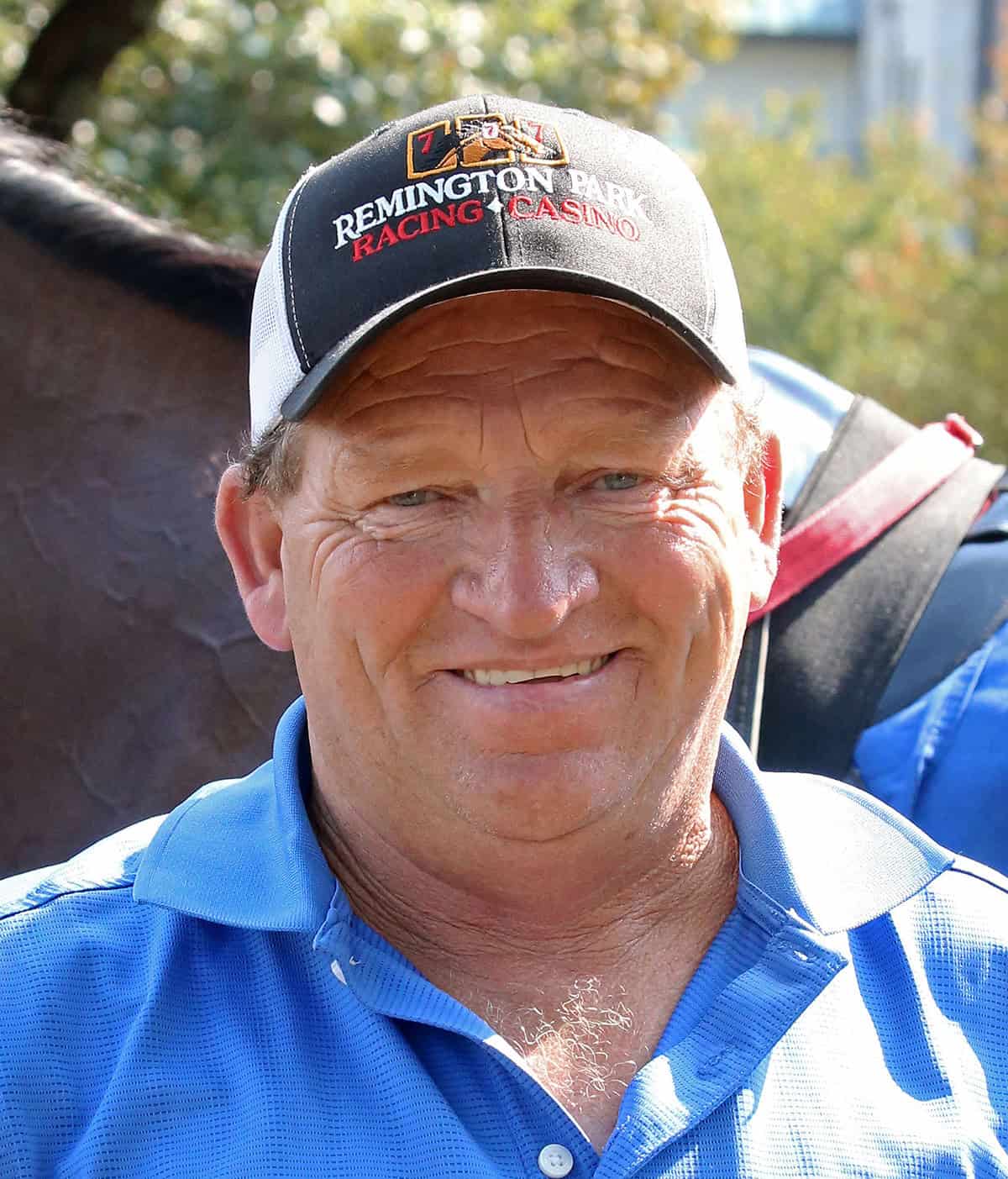 Headshot of trainer Steve Manley. Manley smiles for the camera, wearing a Remington Park Casino & Racing hat, while standing in front of one of his horses. (Photo: ©Coady Media)