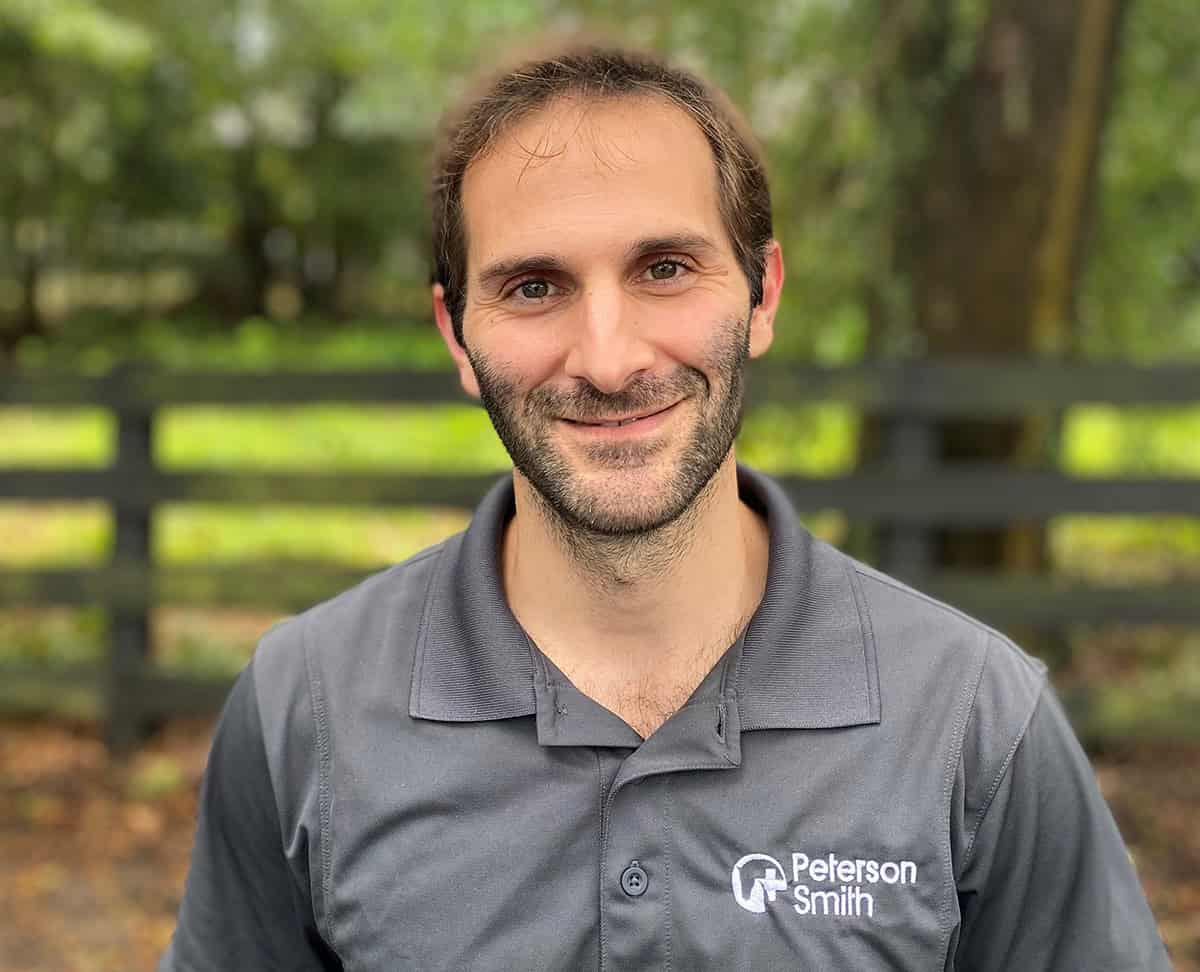 Dr. Marcos Perez, LV, MSc, DACVS-LA. Dr. Marcos Perez is a boarded surgeon at Peterson Smith Equine Hospital + Complete Care in Ocala, Florida, smiles for a photo. He stands in front of a wooden pasture fence, with the background blurred out behind him. (Photo: ©Peterson Smith Equine Hospital + Complete Care)