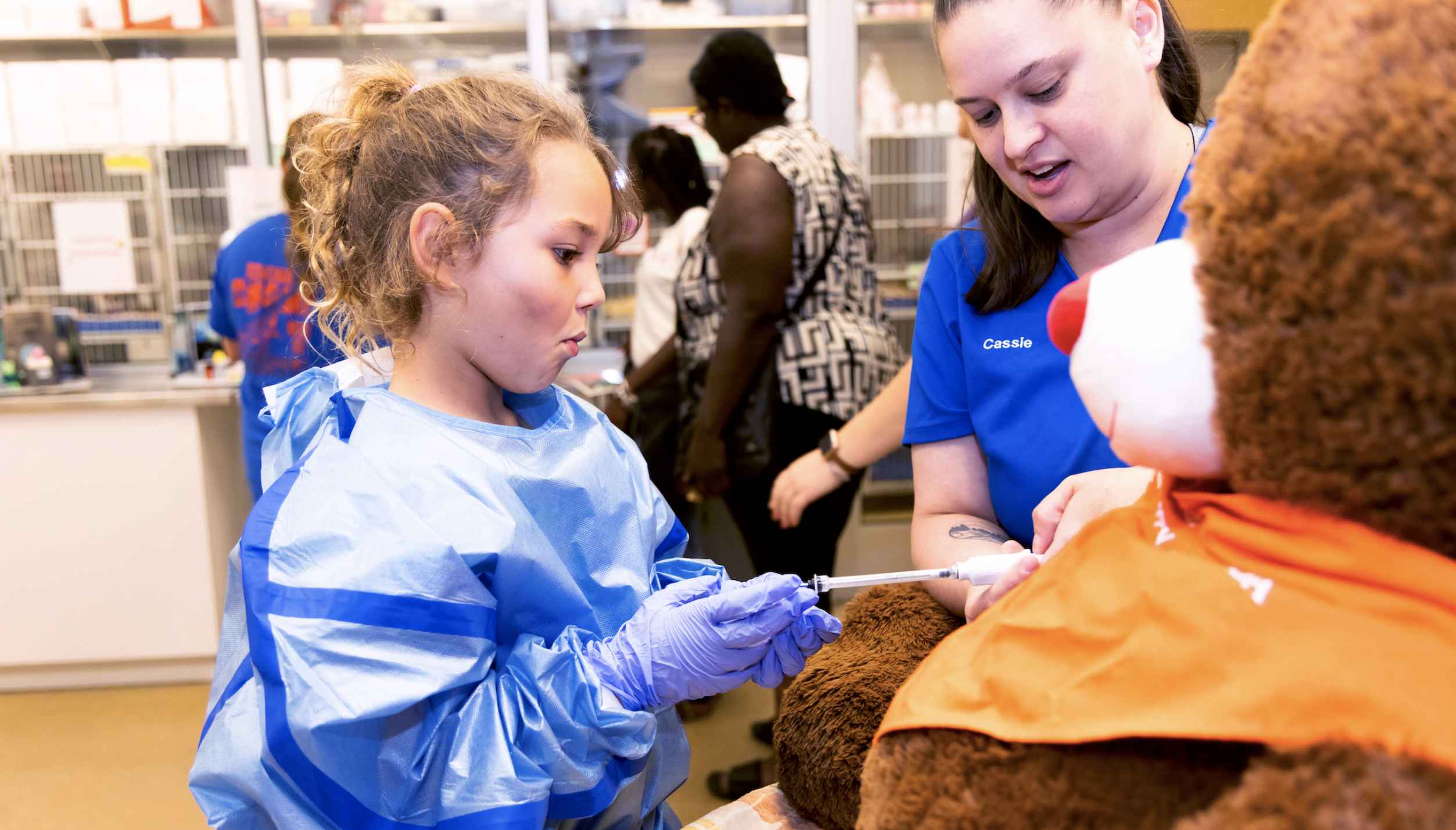 A little girl practices giving a shot to a teddy bear at a veterinarian clinic, dressed in scrubs, while one of the clinic officials guides her. (Photo Courtesy: UF Health News)