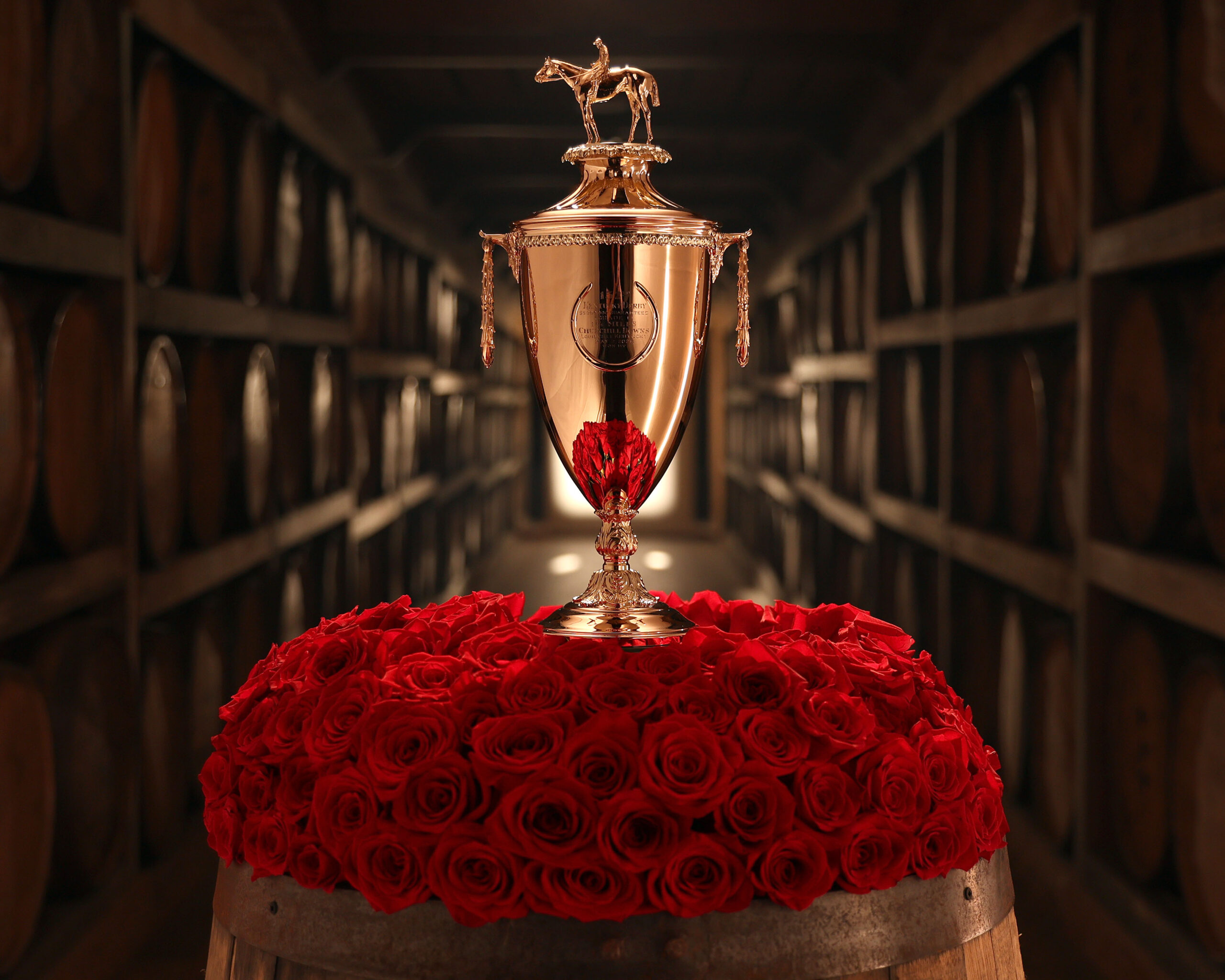 The golden trophy for the 2026 Kentucky Derby is showcased on top of a pile of bright red roses. Light hits the trophy, illuminating the surrounding area. Wooden whiskey barrels stretch out into the blurred background behind the trophy. (Photo: ©Kurtis Coady / Coady Media)