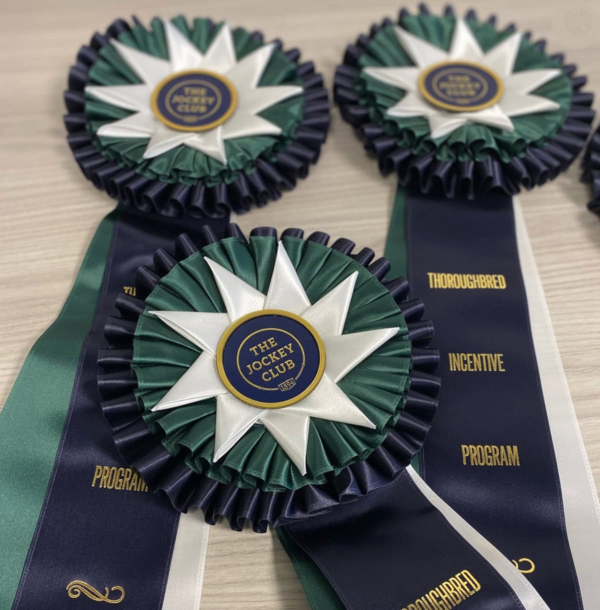 A collection of three dark green and blue ribbons are layered over each other on a table. In the middle of the ribbon’s rosettes, all capitalized text reads, “The Jockey Club.” Photo courtesy of TIP’s website, tjctip.com.