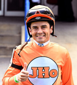 Headshot of jockey Sheldon Russell, smiling for the camera and offering a thumbs up, while dressed in orange silks. (Photo: ©Jim McCue)