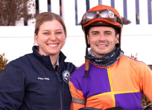 Brittany (left) and Sheldon (right) Russell smile for the camera. Brittany wears a Breeders' Cup jacket, and Sheldon wears bright orange and purple jockey silks. (Photo: ©Jim McCue/Maryland Jockey Club)