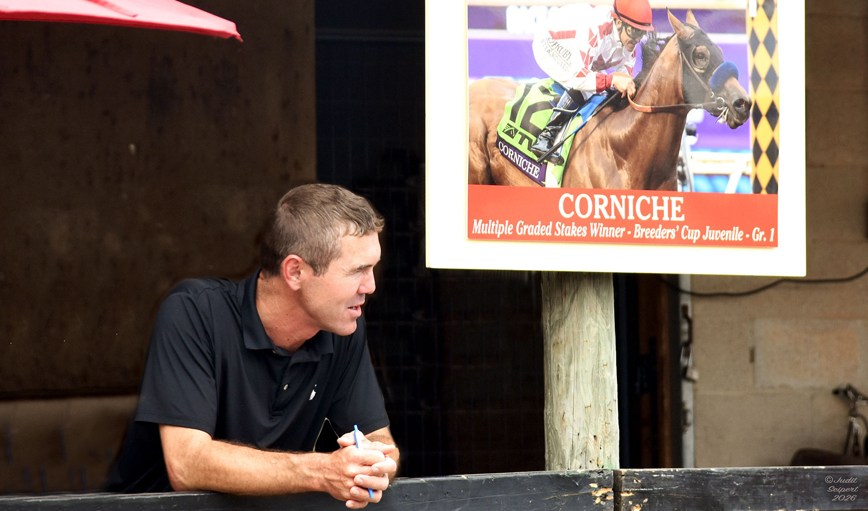 Tristan de Meric leans forward against a rail in front of horse stalls, holding a pen in his hands while viewing (presumably) horses passing by. He’s standing next to a photo of a horse that graduated from his program, Corniche. The sign further describes Corniche as a Multiple Graded Stakes winner and winner of the 2021 Breeders Cup Juvenile (G1). (Photo: ©Judit Seipert)