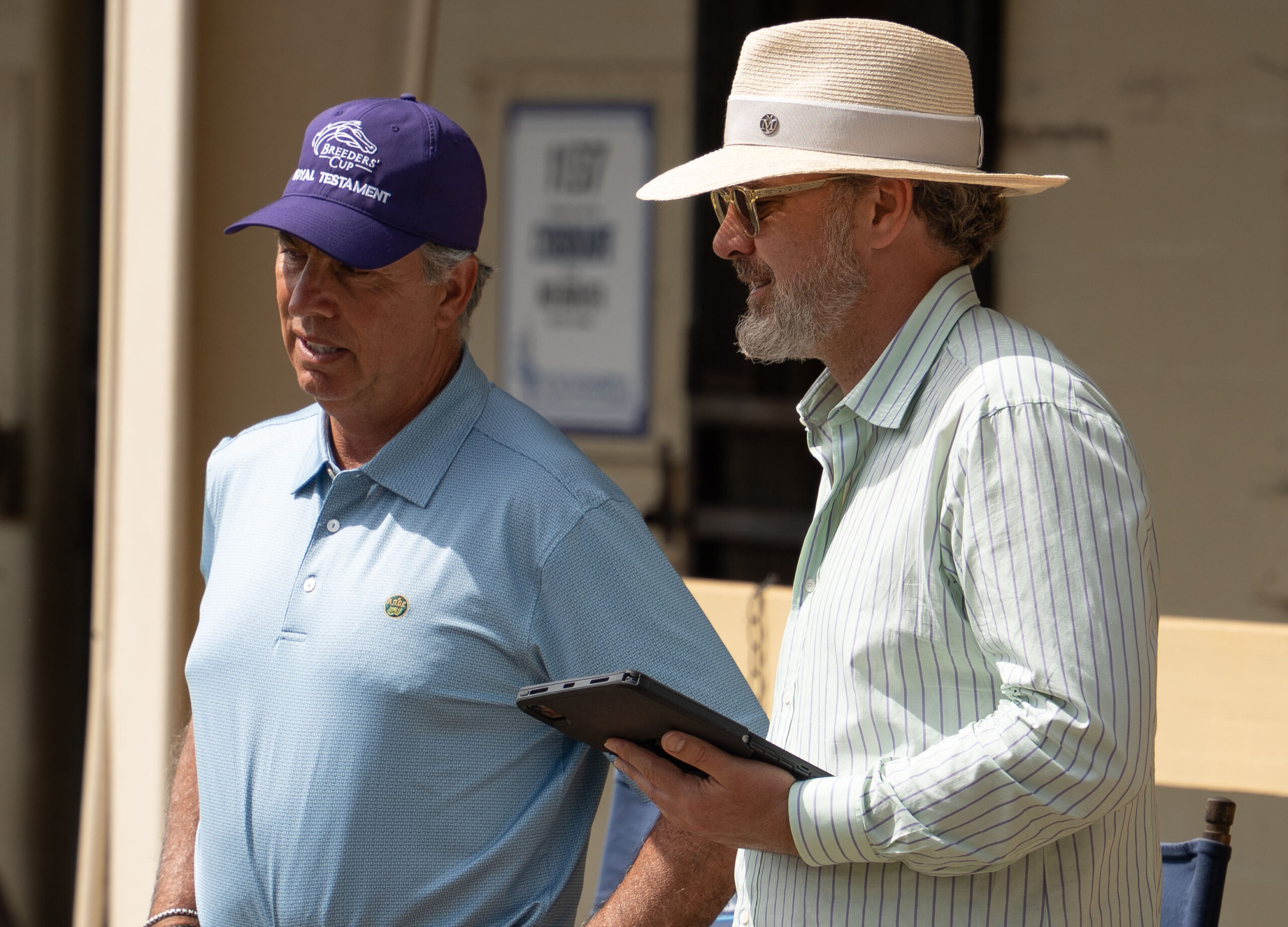 Cropped photo of two people standing outside the Ocala Breeders’ Sales Company sale barns. The man on the left wears a purple Breeders’ Cup hat, while the man on the right of the photo holds an iPad. The two are in conversation, presumably looking at a horse to purchase. (Photo: ©OBS Photo)