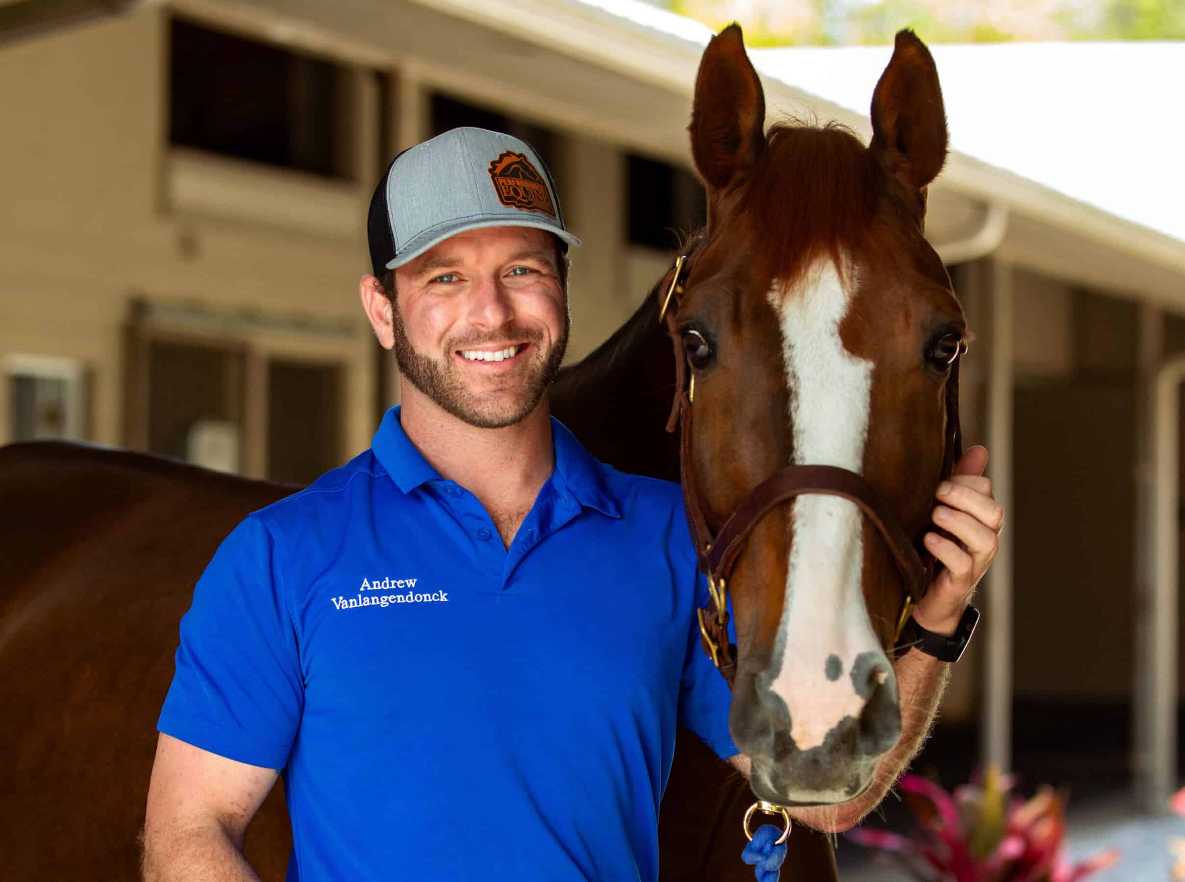 Photo of Andrew Vanlangendonck smiling for a photo with a chestnut horse with a blaze standing next to him. The horse’s ears are pricked as they look towards the camera. Together, they stand in the middle of a barn aisle.
