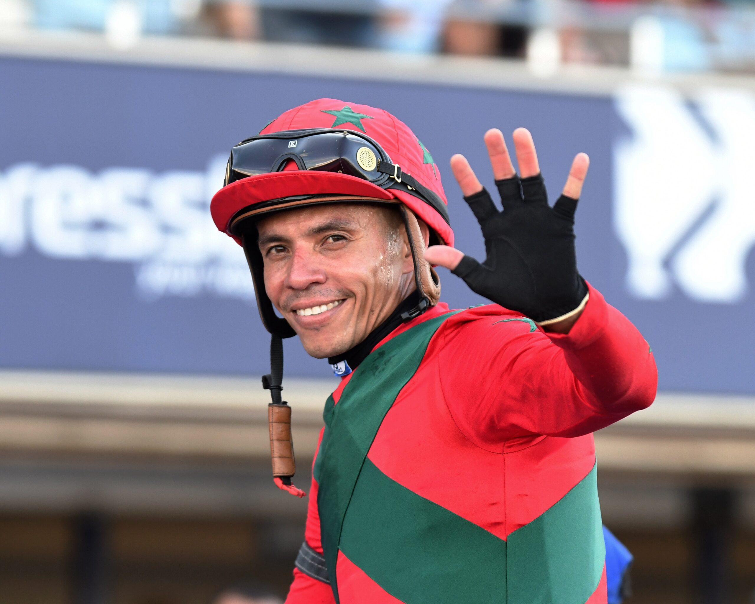 Closely cropped photo of jockey Emisael Jaramillo smiling waving to the camera while riding a horse (out of frame) off the track post win. (Photo: ©Ryan Thompson)