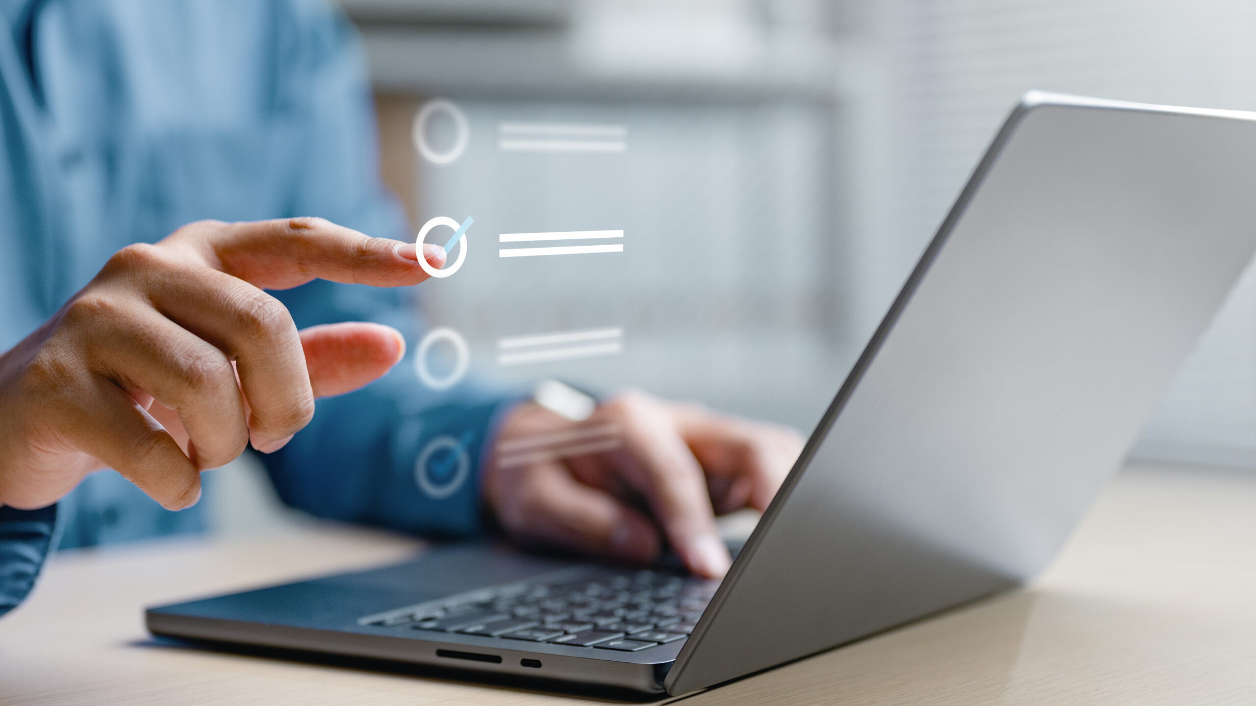 A person’s left hand rests on a laptop’s keyboard, while their right hand is raised, checking off an item on a floating digital list that acts as an extension of the laptop. (Photo: ©Nutthaseth Vanchaichana)