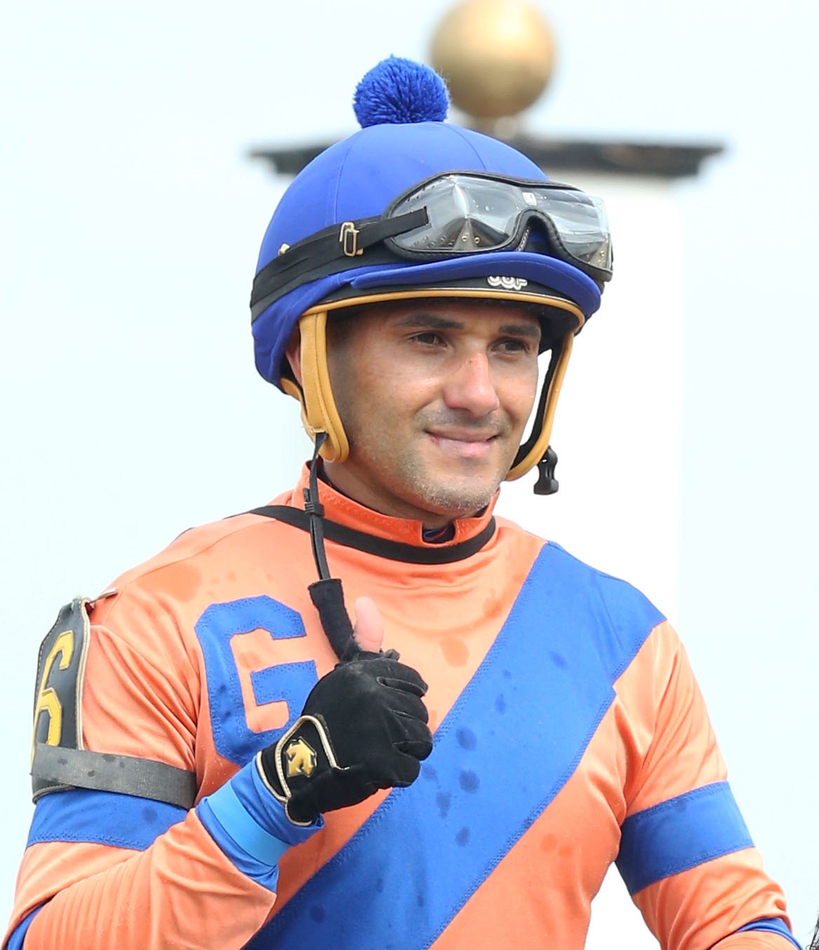 Closely cropped headshot of jockey Orlando Mojica giving a thumbs up to the camera while (presumably) riding a horse post race. (Photo: ©Coady Media)