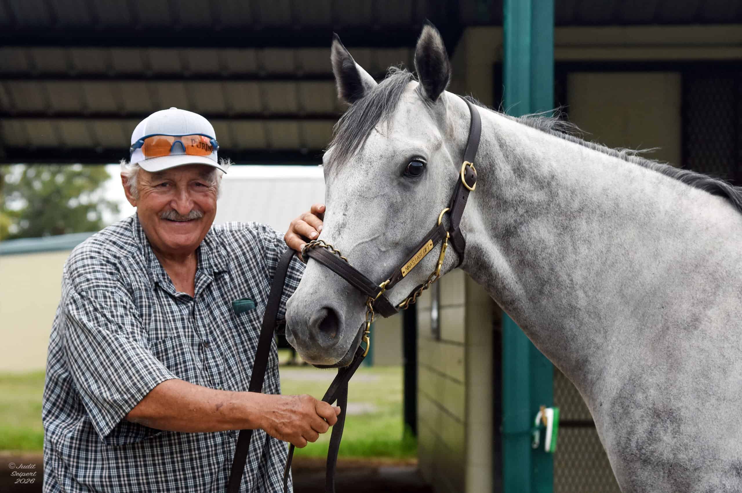 Headshot of Max Ubide smiling at the camera while petting Nina’s Last Gift, his gray Florida-bred Thoroughbred homebred filly. The pair pose for the photo by the barns of the OBS Sale grounds. (Photo: ©Judit Seipert)