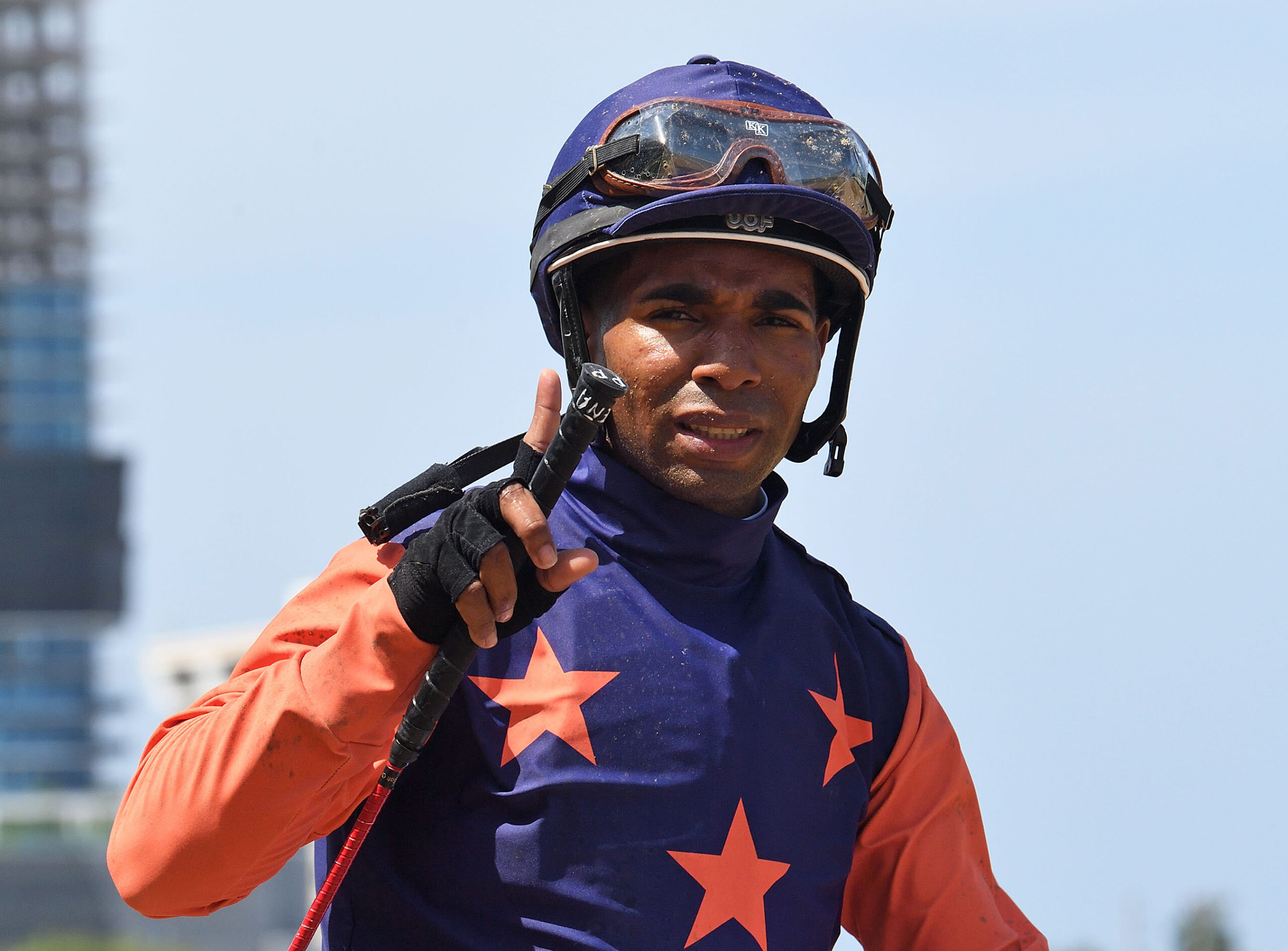 Closely cropped headshot of jockey Pedro Pena, who looks to the camera holding up a finger to signal and celebrate his most recent win as he rides off the track. (Photo: ©Ryan Thompson)
