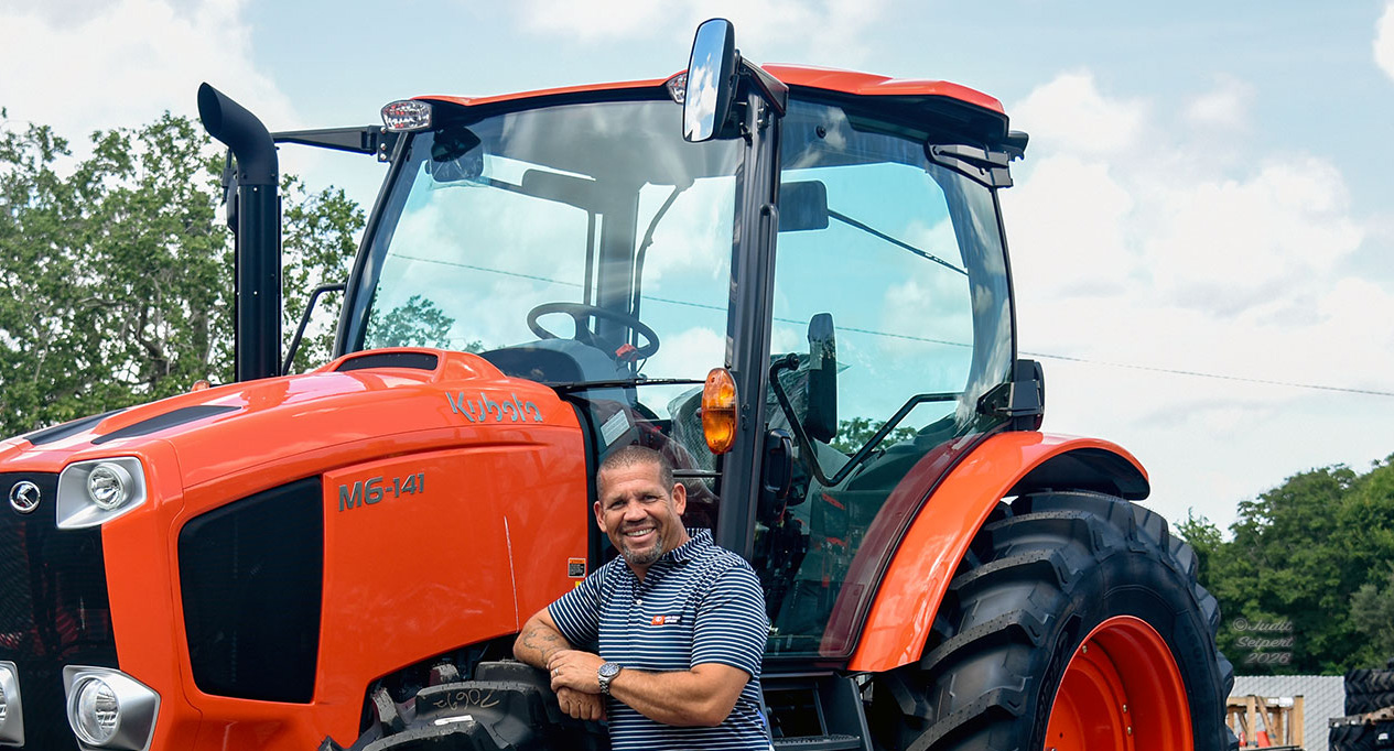 Isaiah Webb, general manager of Florida Coast Equipment’s Ocala Kubota dealership, leans against the tire of an orange Kubota tractor. The photo is cropped from the waist up. (Photo: ©Judit Seipert)