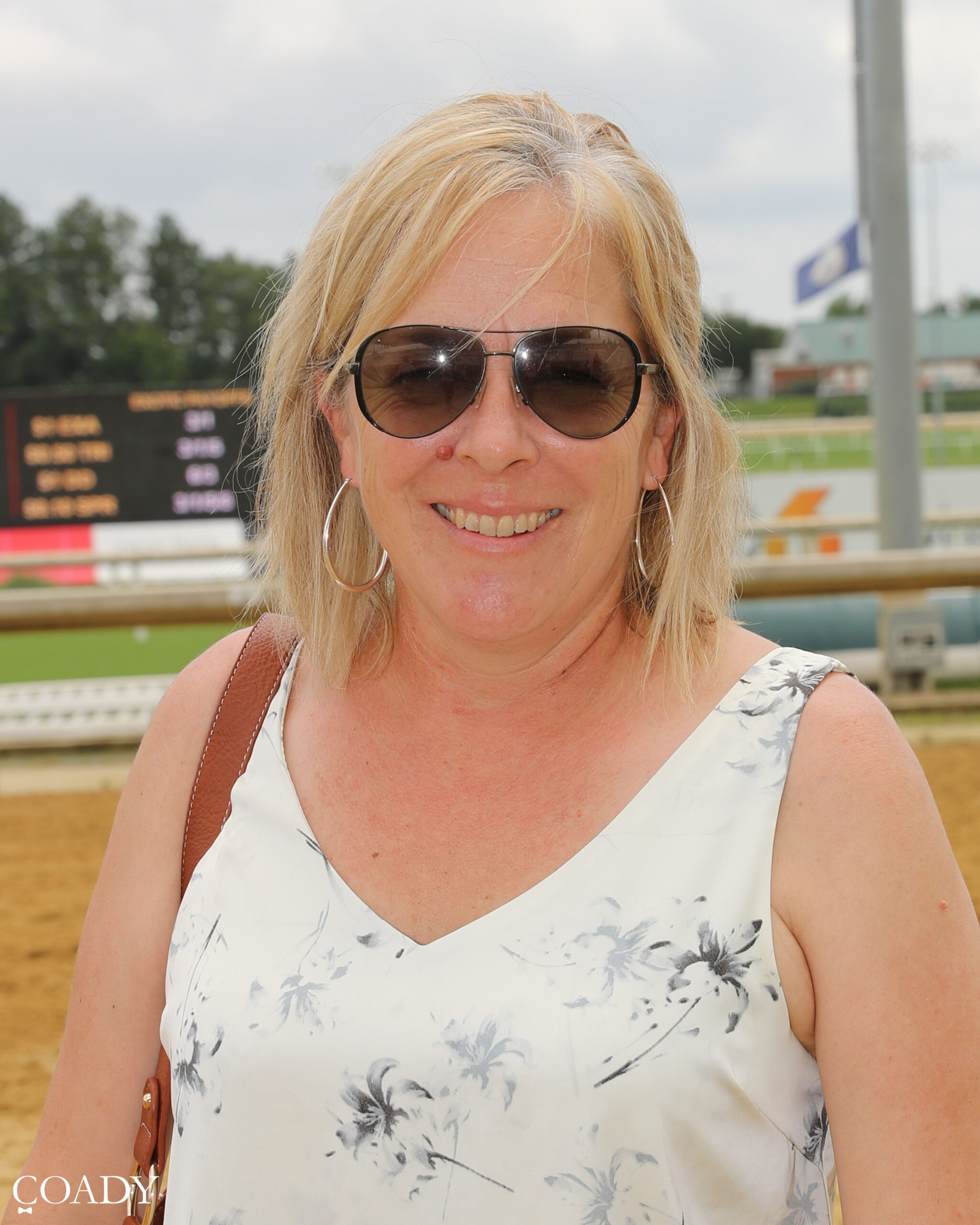 Headshot of trainer Maria Bowersock wearing sunglasses and smiling for the camera next to the racetrack rail. (Photo: ©Coady Media)