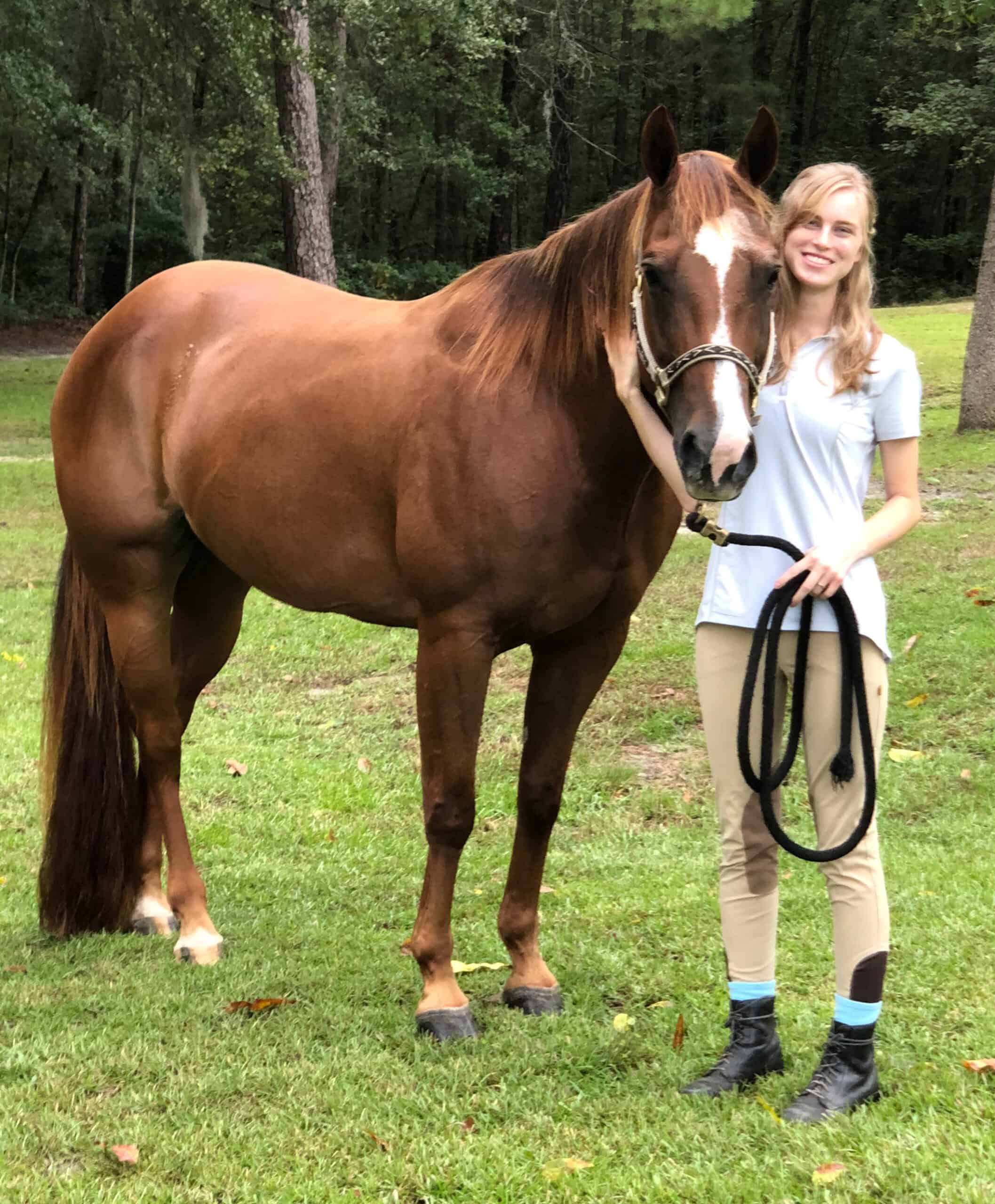 Sorrel Quarter Horse mare, Pass the Paprika, and Jayne Chapman, stand together for a photo. Paprika’s ears are pricked as Jayne holds her lead. Trees line the background of the photo. (Photo courtesy Jayne Chapman)