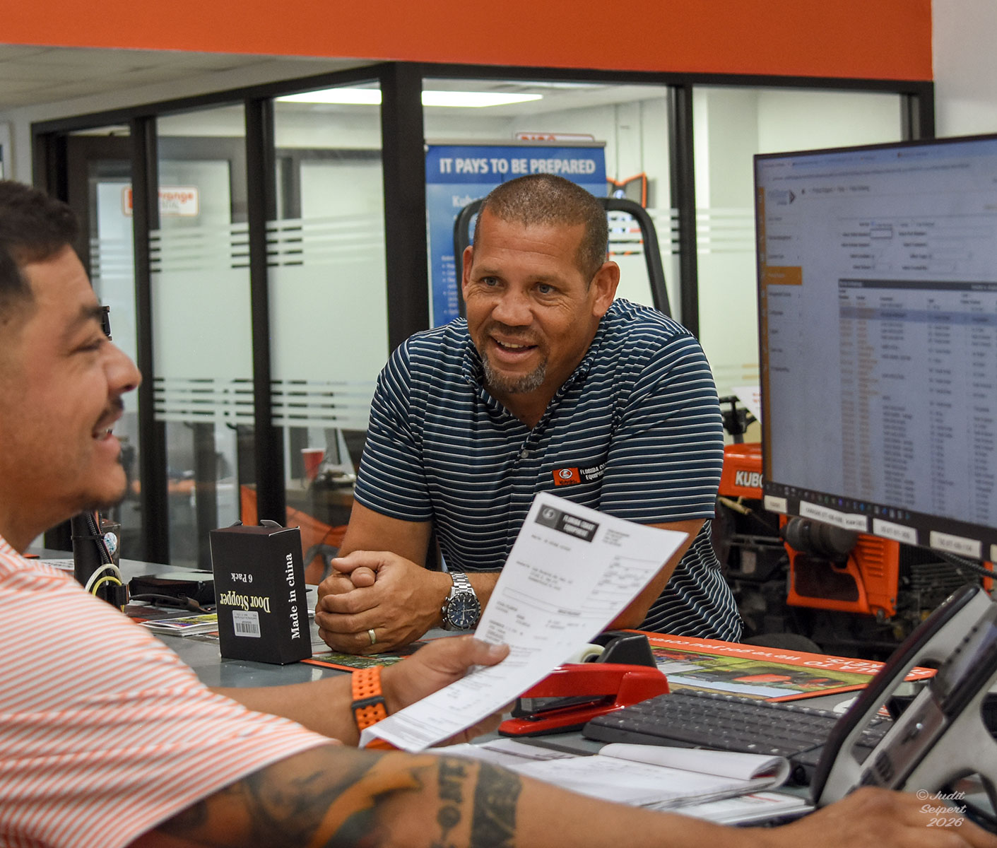 Isaiah Webb leans across the counter of Florida Coast Equipment’s Ocala Kubota dealership, chatting with a fellow employee working at the front desk’s computer. (Photo: ©Judit Seipert)