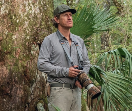 Carlton Ward Jr. stands looking into the distance, camera in hand, against a backdrop of dense trees. (Photo Courtesy: HHF Website)