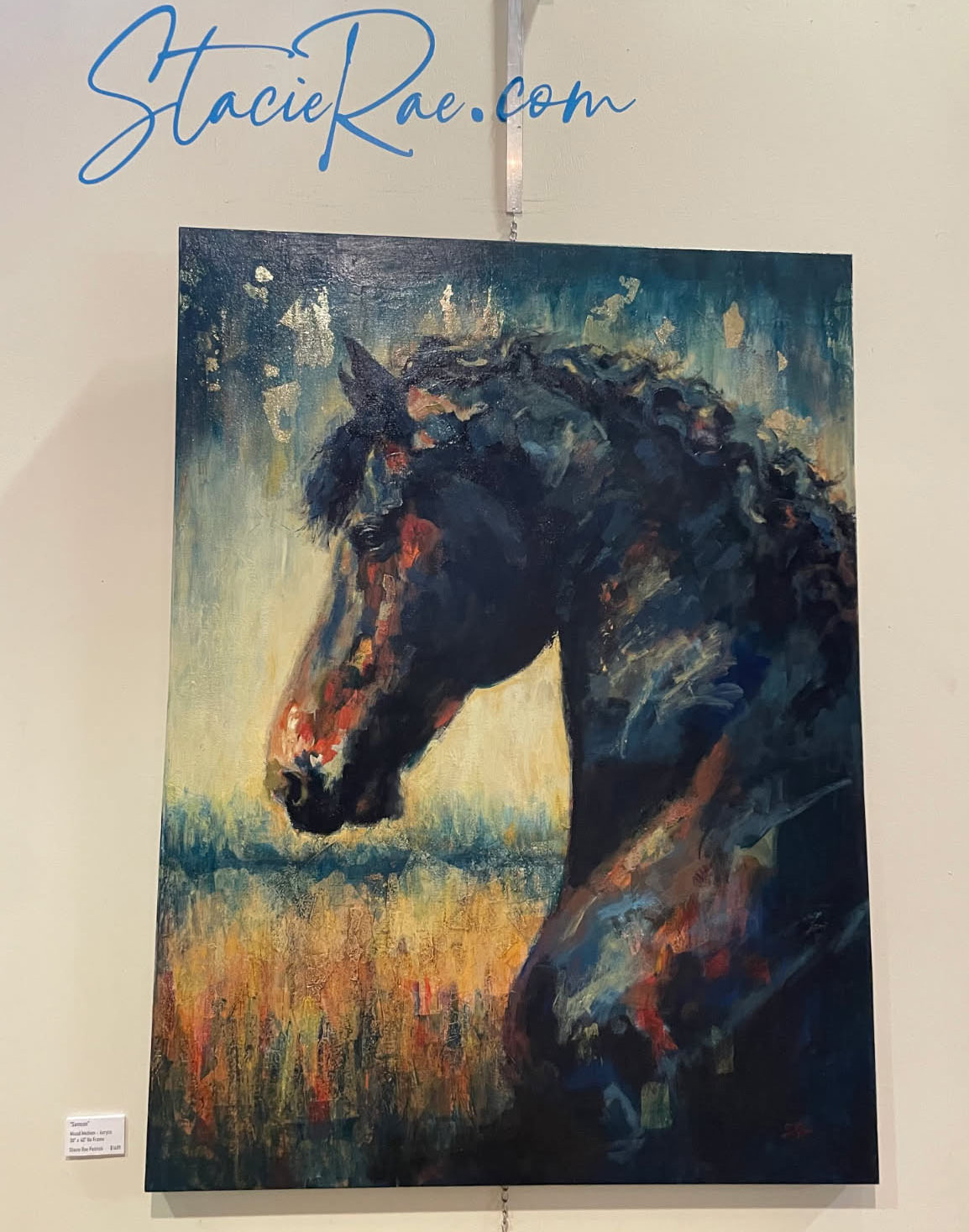 Photograph taken of a painting hanging in a gallery, with hand scripted text written above the painting in blue on the wall. The text reads, “StacieRae.com.” The painting is of a bay horse’s head, accomplished with noticeable brushstrokes that give an impressionistic appearance.