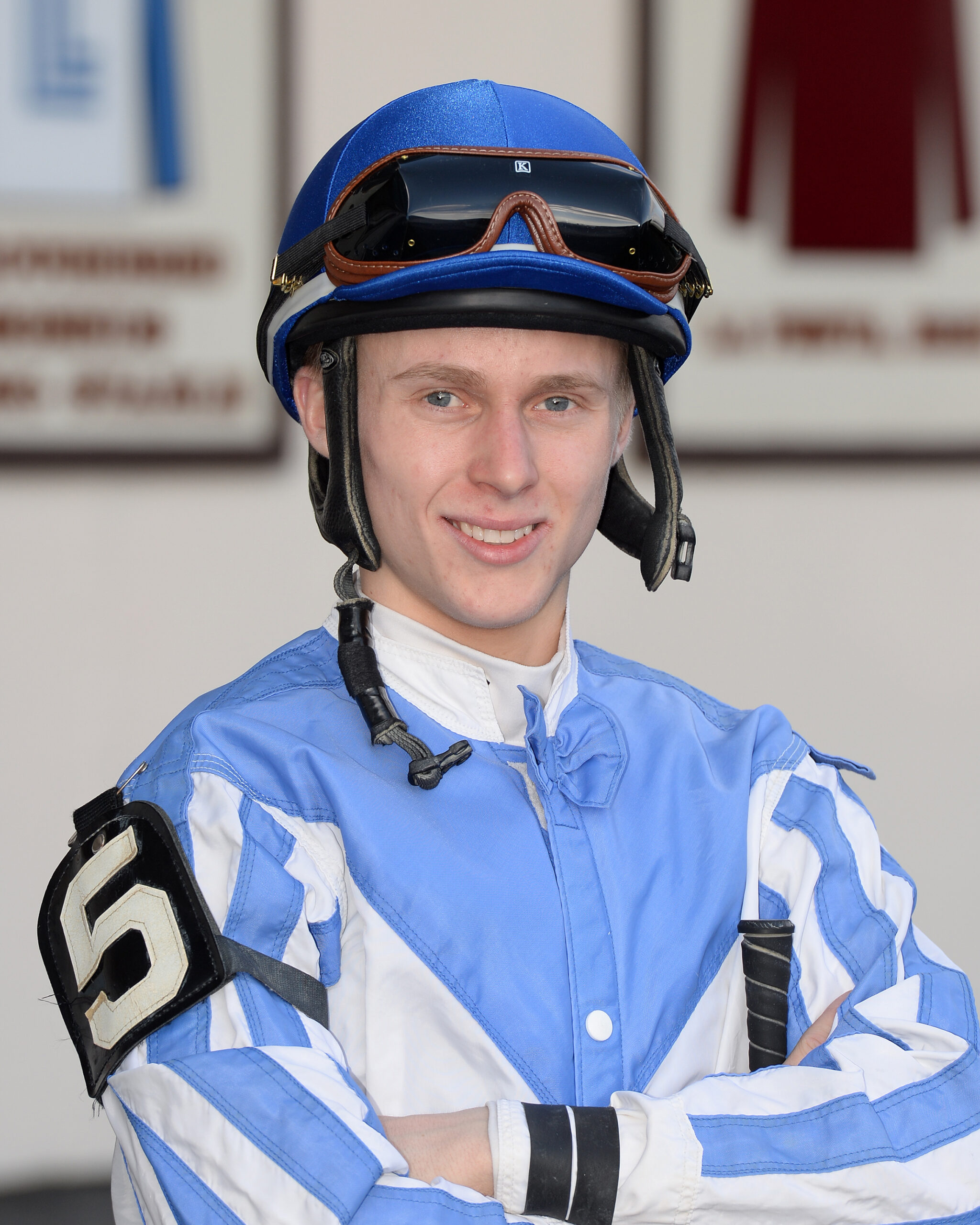 Headshot of jockey Dylan Davis dressed in blue and white silks. His arms are crossed as he stands posed, smiling for the camera. (Photo: ©Coglianese)