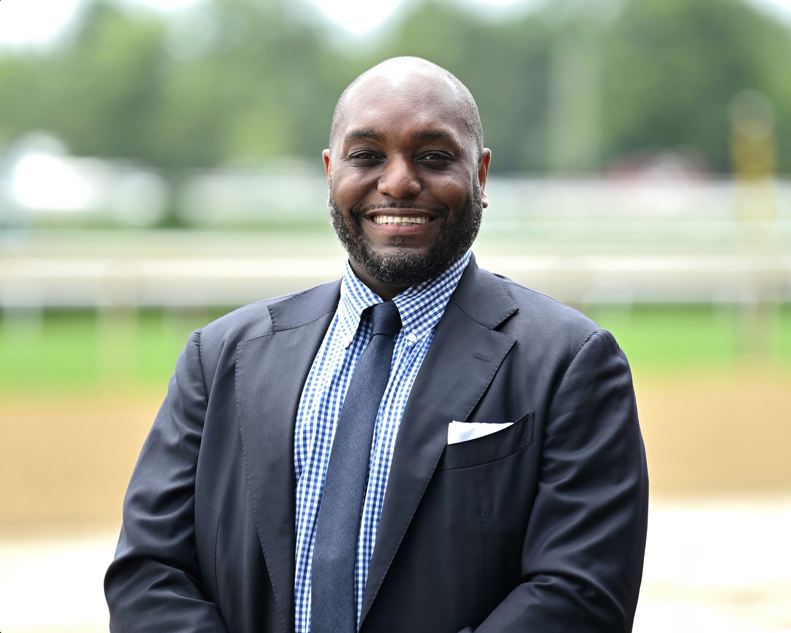 Professional headshot of Najja Thompson smiling for the camera while standing in front of a Thoroughbred racetrack, which is blurred into the background. (Photo Courtesy: The Jockey Club)