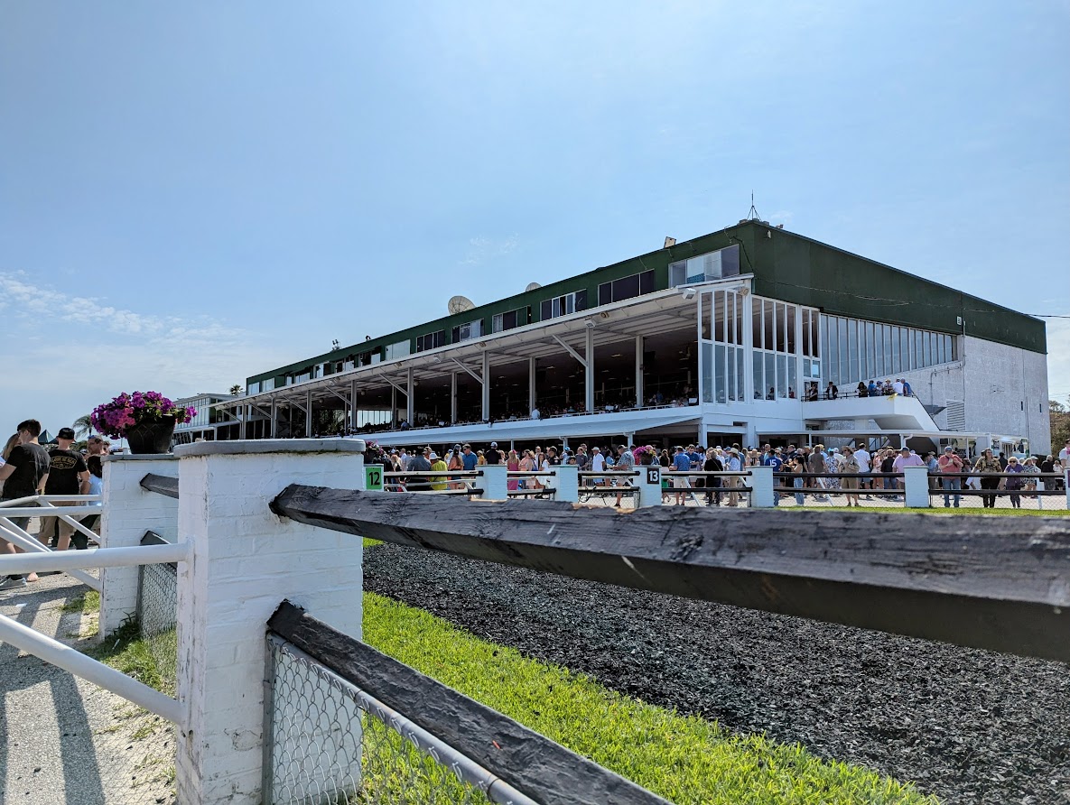 Photograph taken by the paddock rail in front of the grandstand at Tampa Bay Downs on a busy race day. The grandstand and surrounding area is bustling with people. (Photo: ©Tammy A. Gantt)