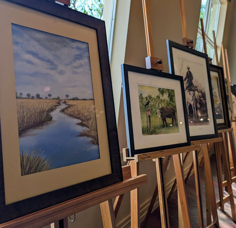 Photograph taken of the art exhibit Ocala Art Group Fall Show titled, Reflections of Farm Life in Marion County. The art shown are all set up on easels set in a neat line against the room’s windows, and are neatly set into black frames. (Photo: ©Tammy Gantt)